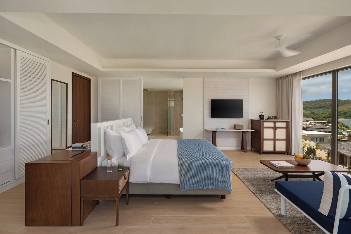 A modern hotel room with a large bed, a desk, a TV, and a balcony with a view.