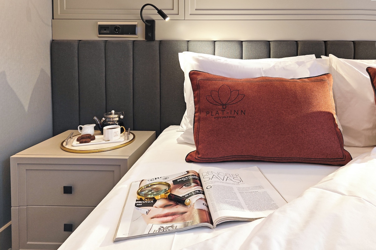 Un letto rifatto con cura, una rivista, un vassoio con teiera e tazze, e un cuscino con il marchio PLAT-INN.