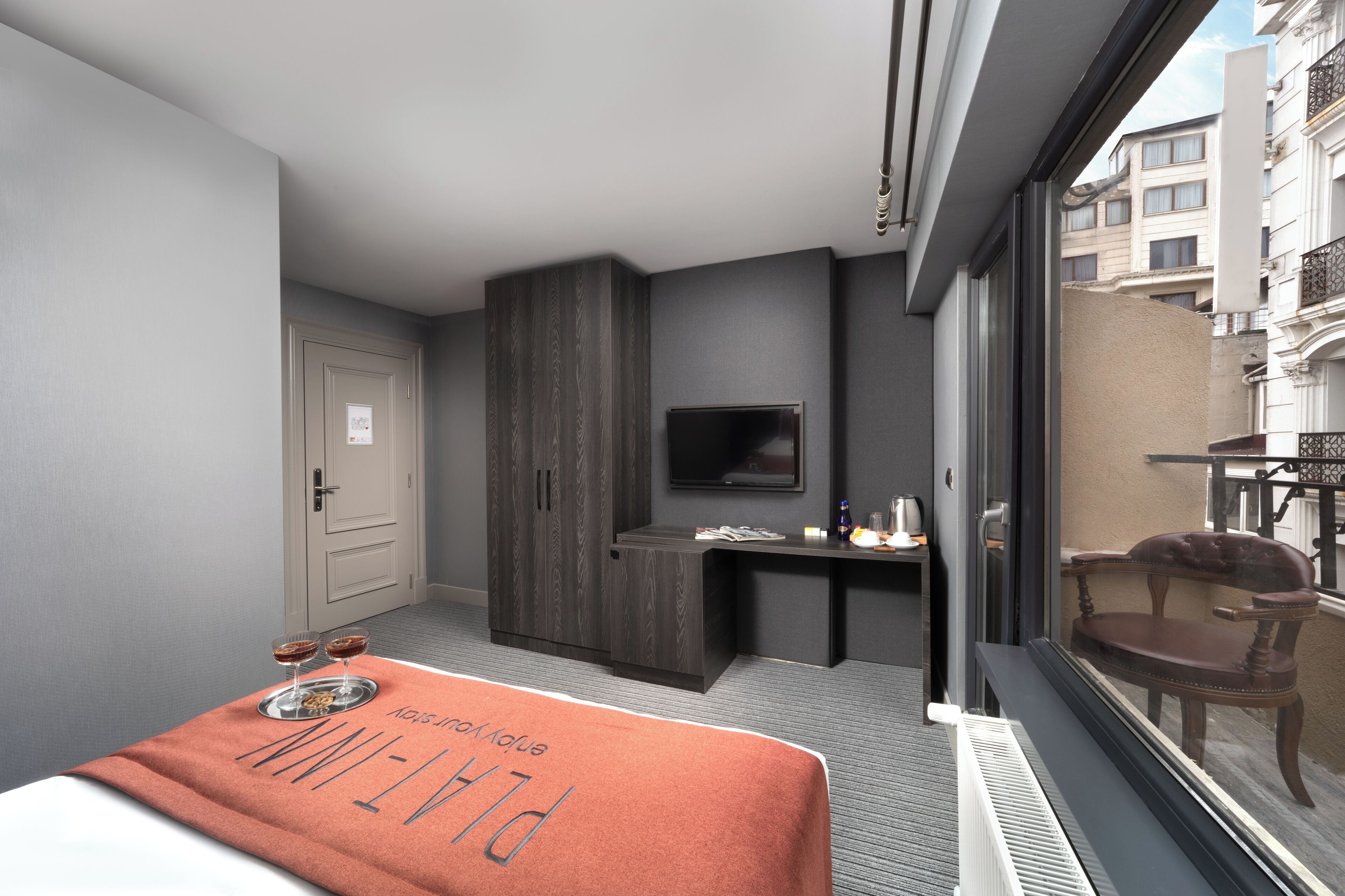 A modern hotel room with a bed, a desk, a TV, and a window with a view of the city.