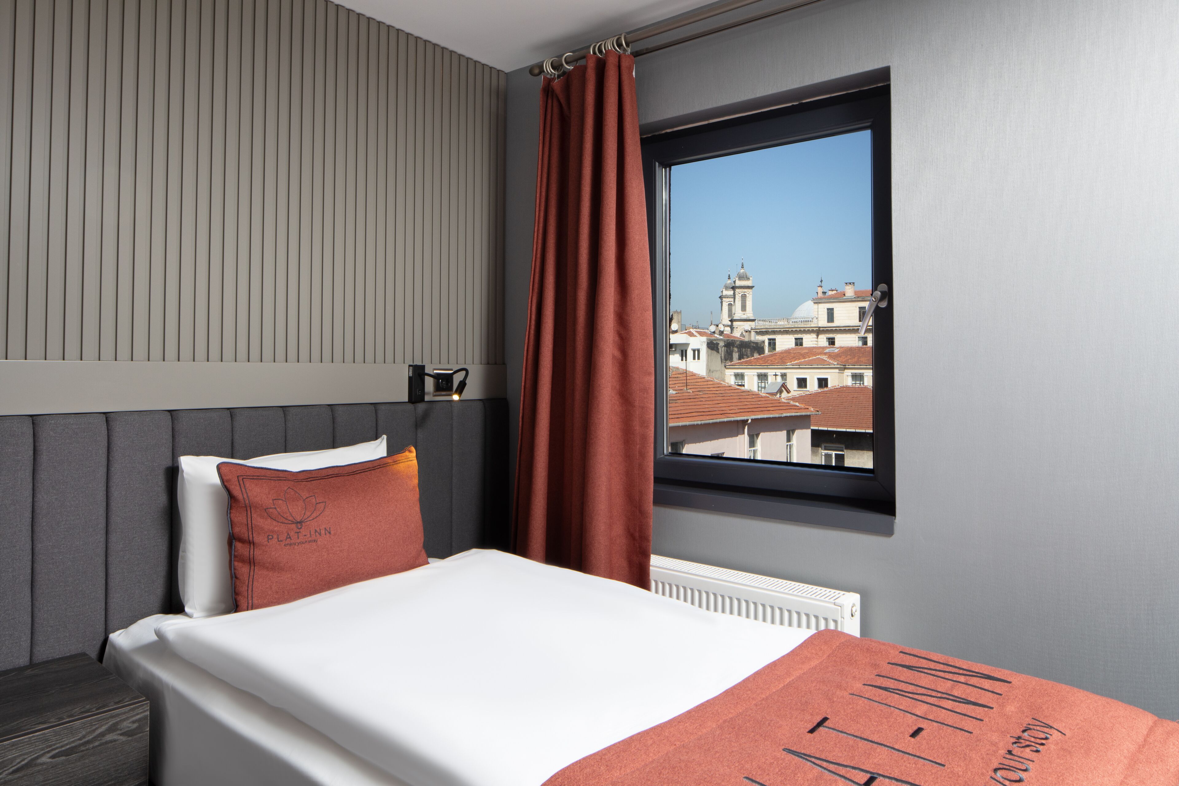 A hotel room with a bed, a window with a view of buildings, a radiator, and a curtain.