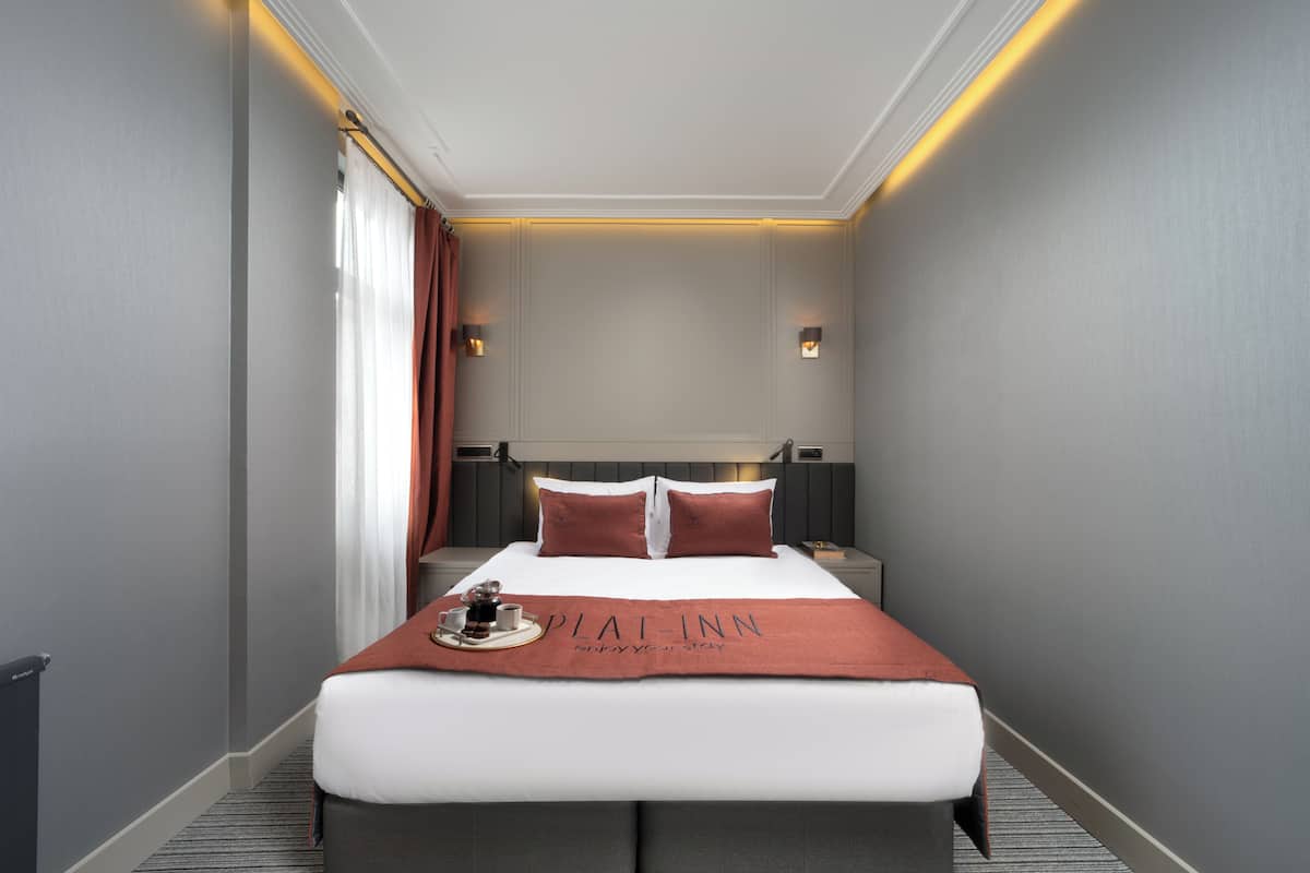 A hotel room with a large bed, a bedside table, a nightstand, a window with curtains, and a wall-mounted light fixture.