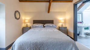 A bedroom with a large bed, two bedside tables with lamps, a wooden ceiling beam, and a view of a bathroom with a sink and mirror. - Buff Lodge - sleeps 4 guests in 2 bedrooms (Near Oxford)