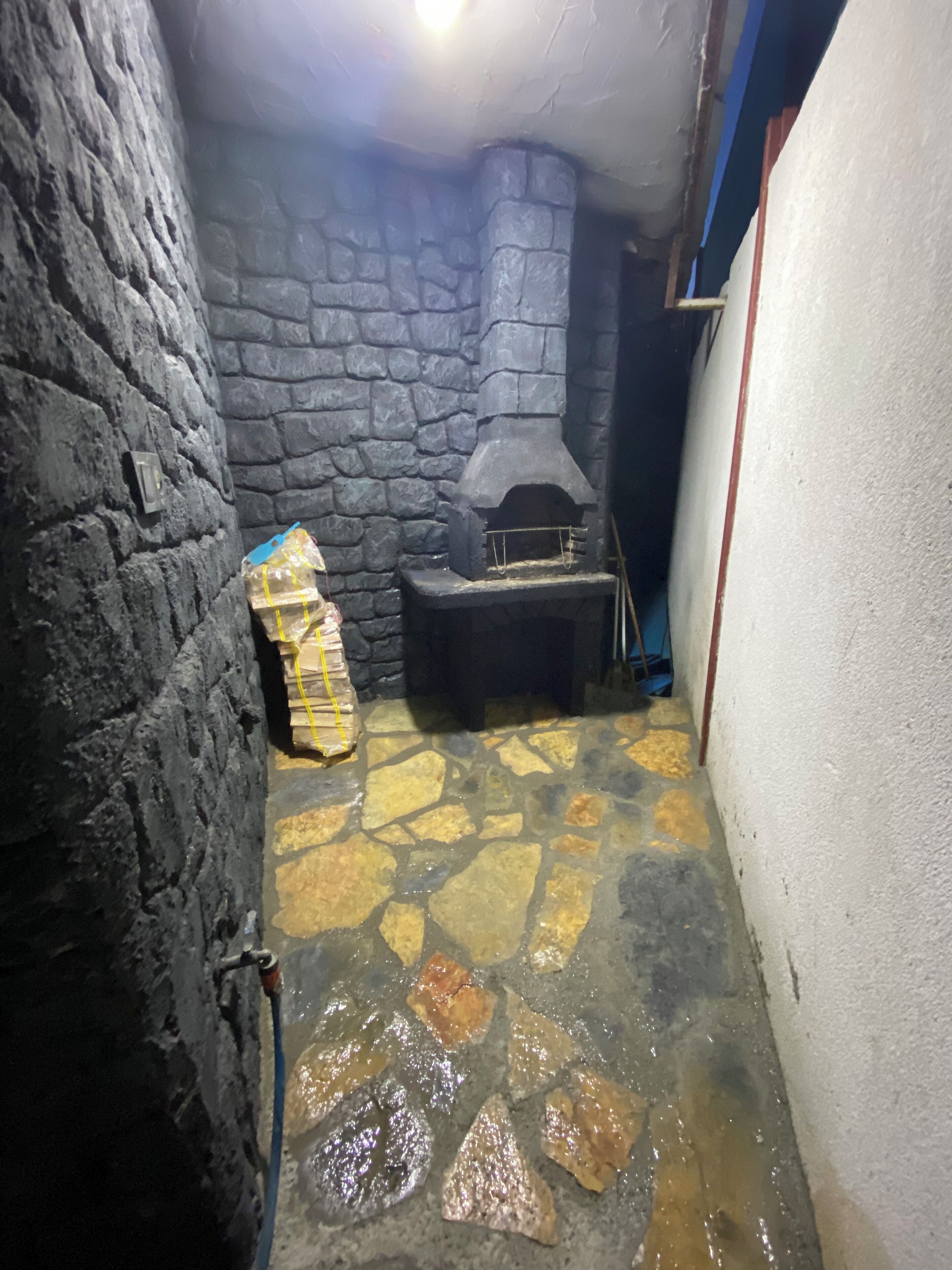 A narrow stone-walled room with a stone floor, a stone fireplace, and a wooden structure on the left.
