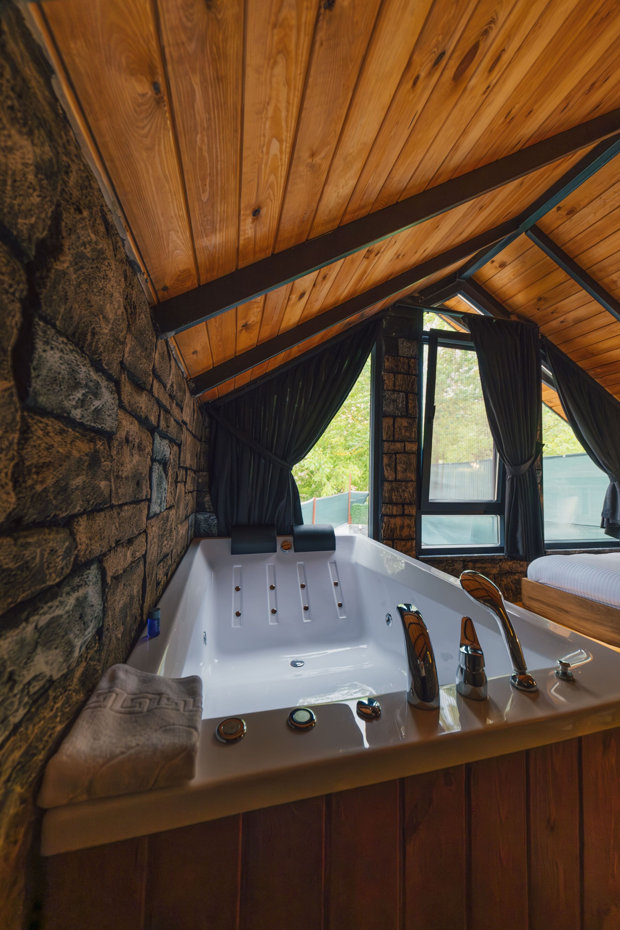 Un plafond en bois, un mur en pierre, une baignoire et une fenêtre avec des rideaux.