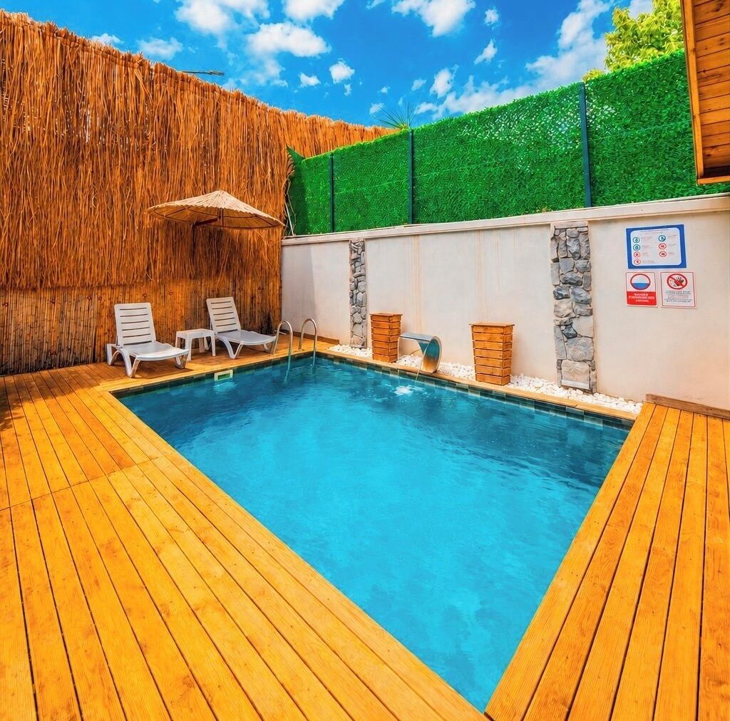 Ein rechteckiger Pool mit Holzterrasse, Liegestühlen und einer Steinmauer mit einer grünen Hecke.