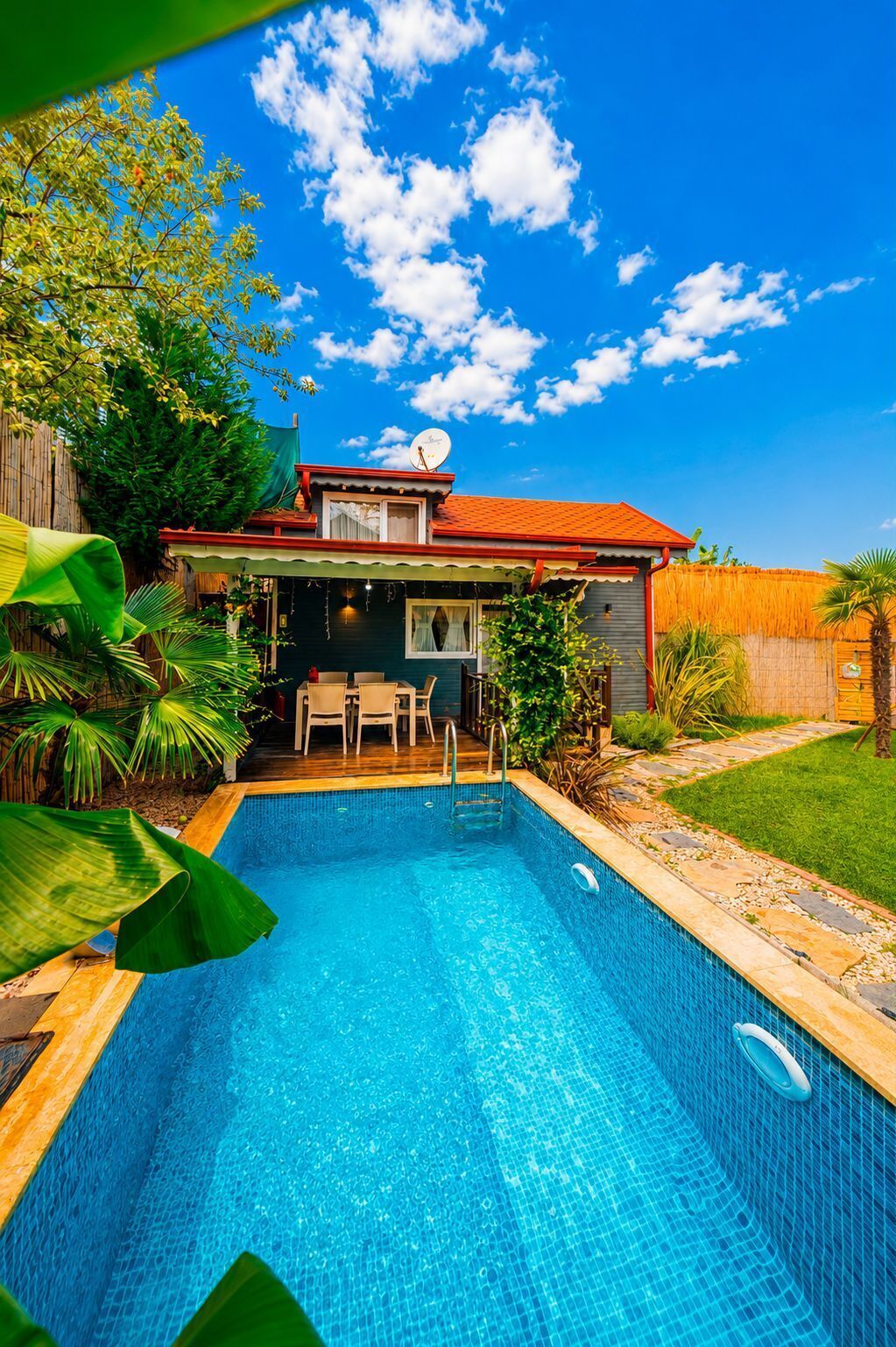A swimming pool with a wooden deck and chairs, surrounded by a fenced yard with a house in the background.
