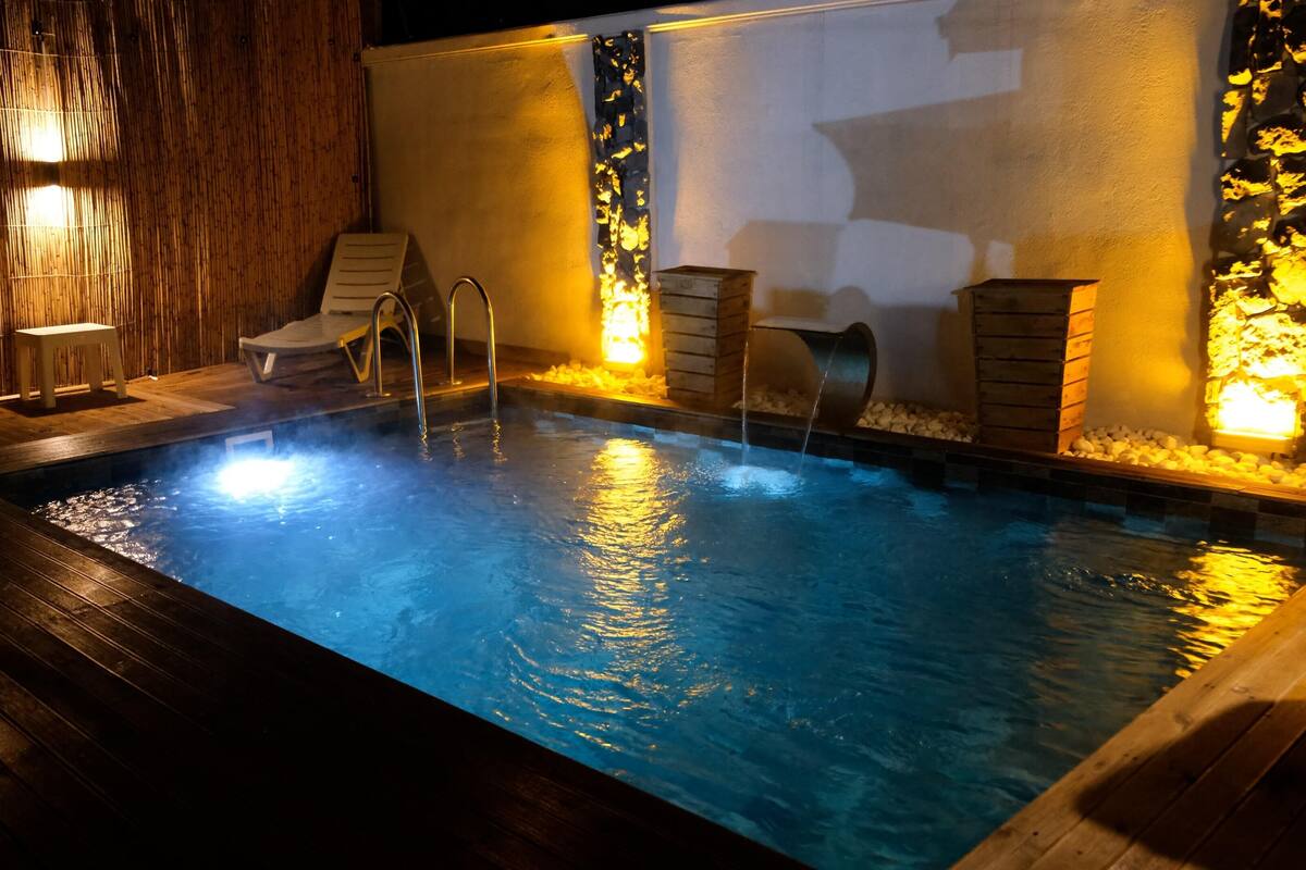 En swimmingpool med ett trädäck och solstolar, omgiven av stenmurar och upplyst av väggmonterade lampor.
