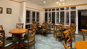 A dining area with wooden tables and chairs, large windows, and a ceiling fan. - Summit Bougainvillea Tea Resort (Kurseong)