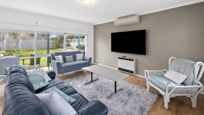 A modern living room with a grey sofa, a white armchair, a coffee table, and a flat-screen TV mounted on the wall. - Ideally located Beach House close to picturesque reserve and beaches (Torquay)