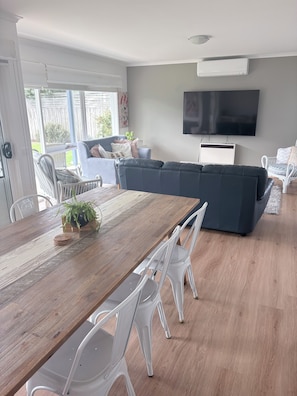 A modern living room with a wooden dining table, white plastic chairs, a grey sofa, and a flat-screen TV mounted on the wall.