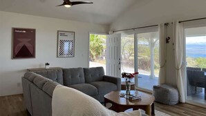 A spacious living room with a grey sofa, a coffee table with books and flowers, a ceiling fan, and a large sliding glass door leading to an outdoor area with a view of the sea. - Hale Lewa ~ Newer Home ~ Ocean Views ~ 30 Day Rent (Waimea)