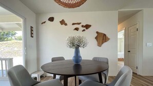 A dining area with a round table, six grey chairs, a blue vase with white flowers, and a wall with wooden and stone decor. - Hale Lewa ~ Newer Home ~ Ocean Views ~ 30 Day Rent (Waimea)