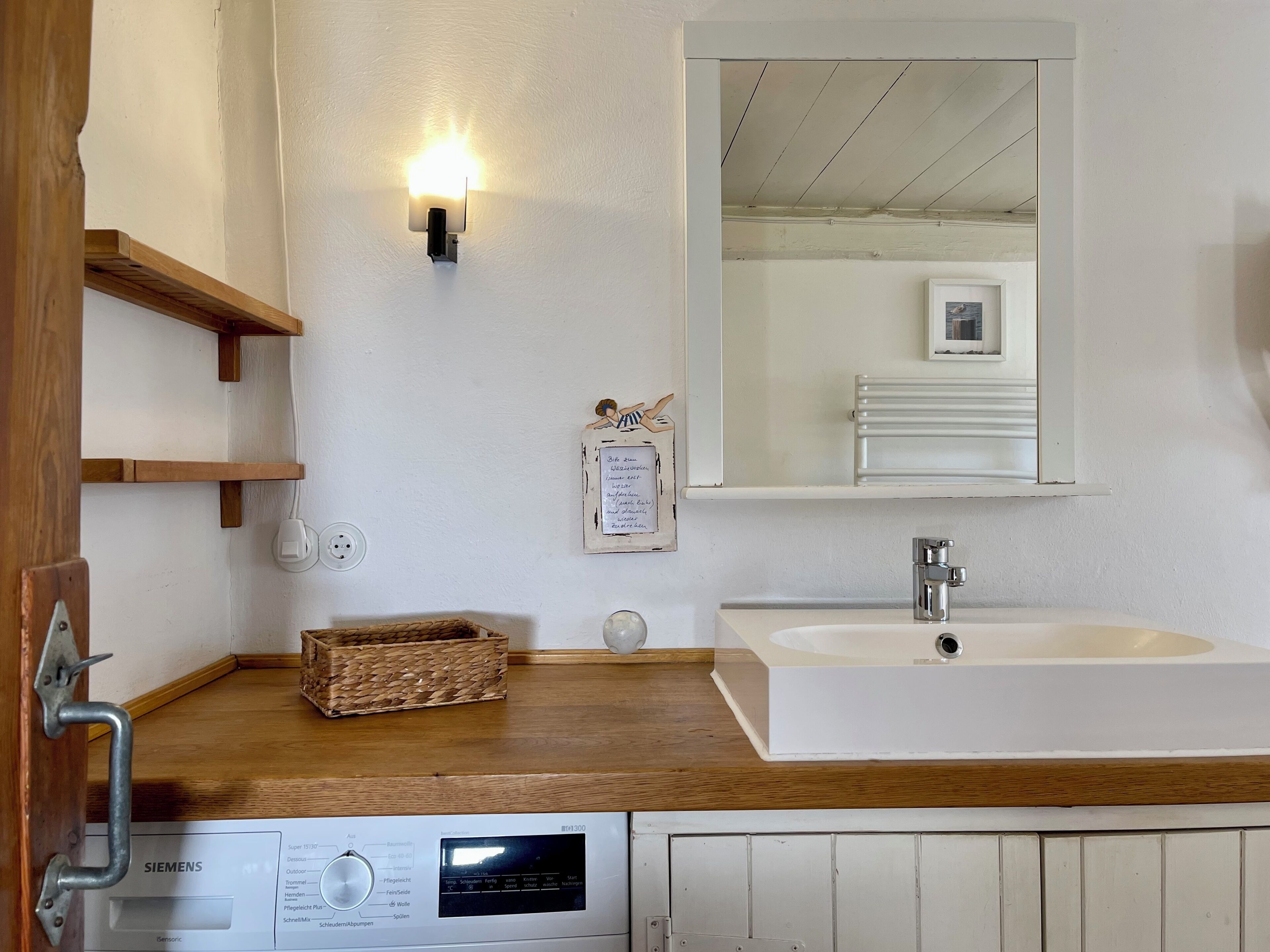 A bathroom with a white sink, wooden shelves, a wall-mounted light, a mirror, and a Siemens device on the counter.