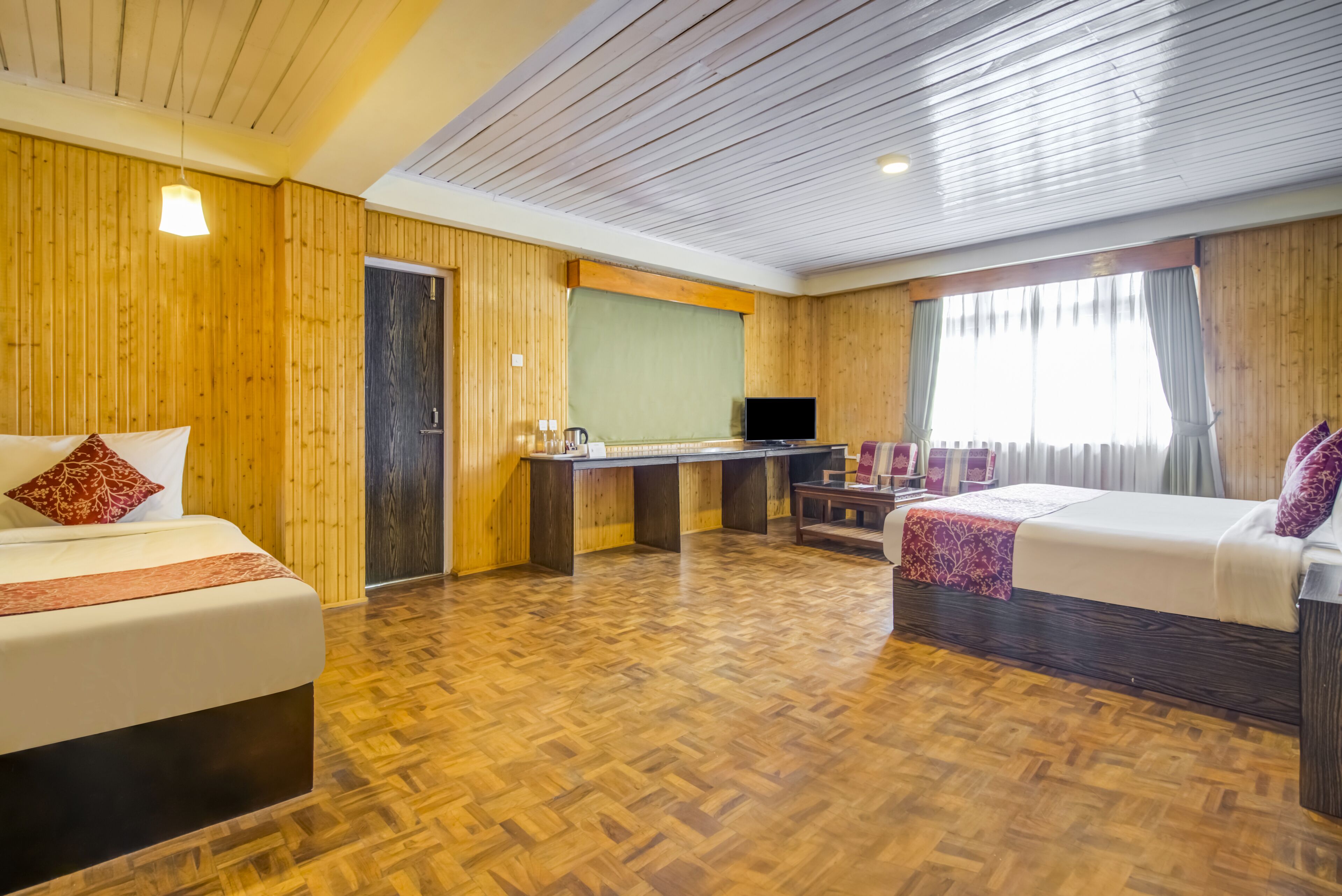 A hotel room with wooden panel walls, a herringbone floor, a bed with a red pillow, a desk with a flat-screen TV, and a window with curtains.
