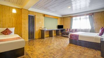 A hotel room with wooden panel walls, a herringbone floor, a bed with a red pillow, a desk with a flat-screen TV, and a window with curtains.