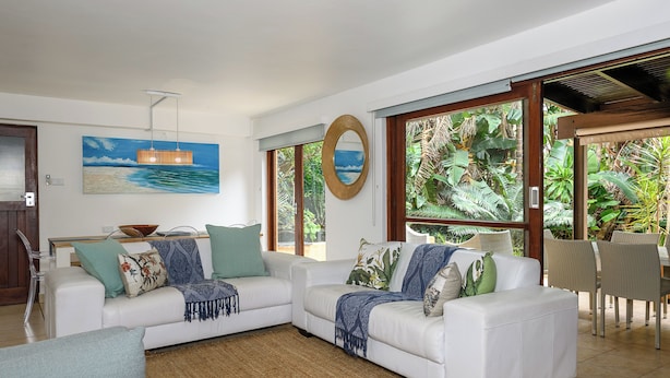A modern living room with a white sofa set, a dining area with wicker chairs, and large windows with tropical views. - San Lameer Villa 2810 (Southbroom)