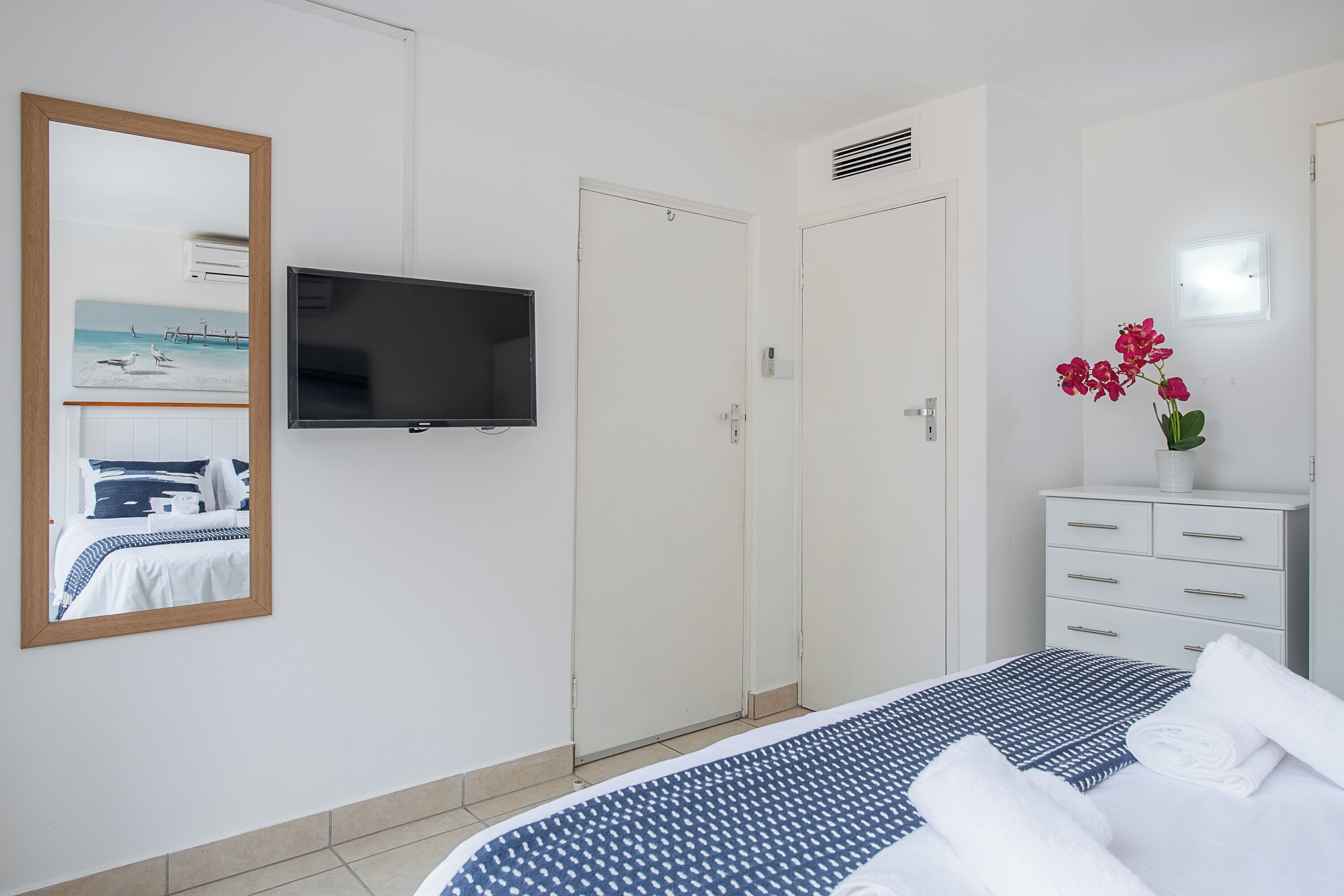 A hotel room with a bed, a TV, a wardrobe, and a mirror reflecting a beach scene.