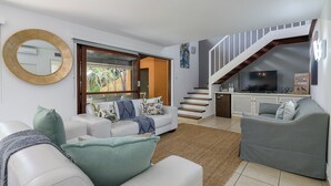 A modern living room with a white sofa, a grey sofa, and a large mirror. There is a staircase leading to an upper level with a TV and artwork. - San Lameer Villa 2810 (Southbroom)