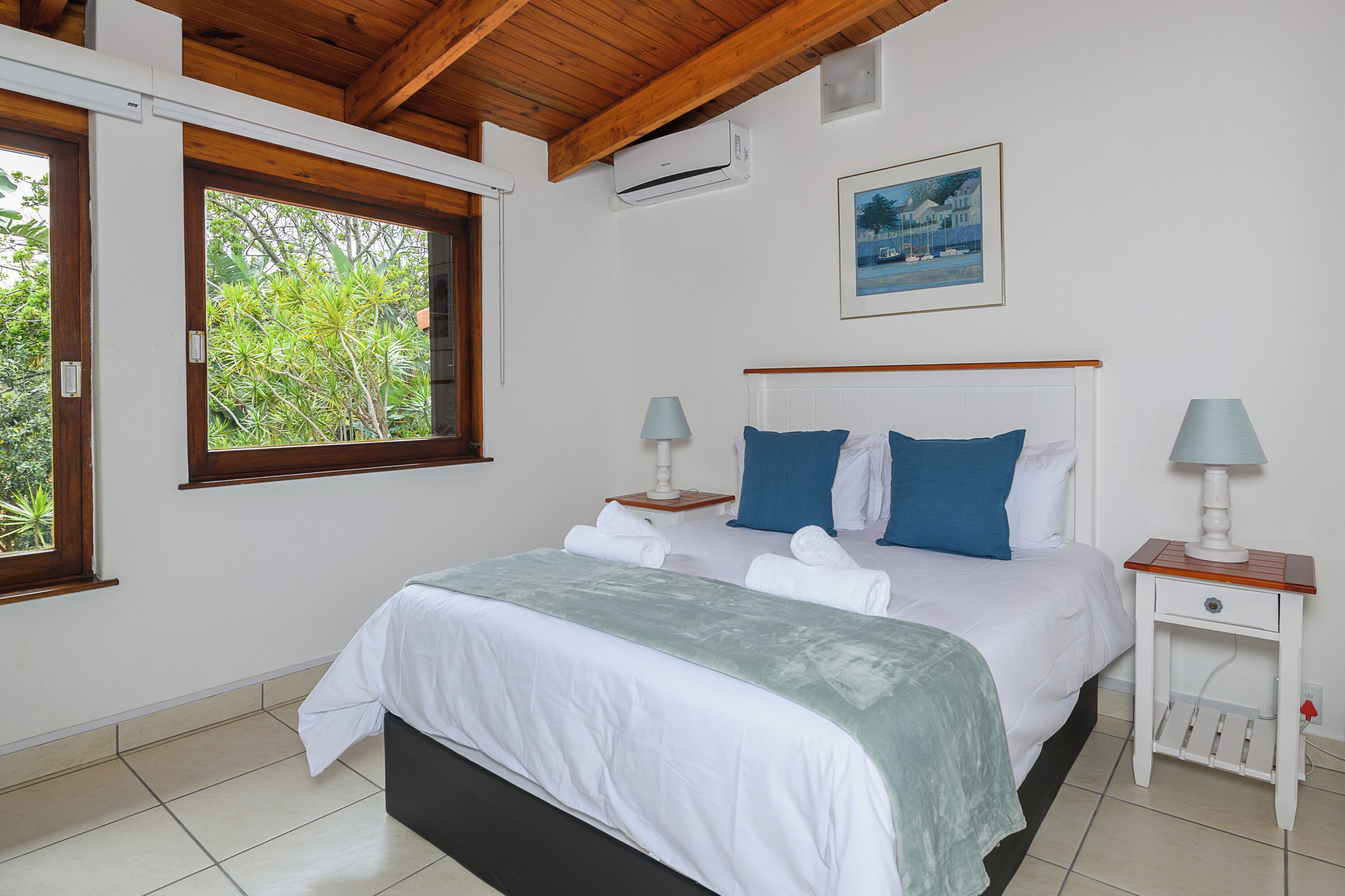 A bedroom with a bed, two bedside tables with lamps, a window with a view of greenery, and an air conditioning unit on the wall.