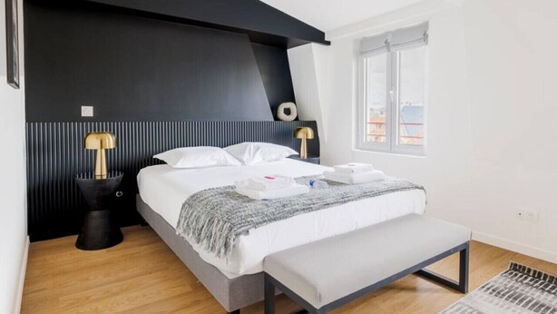 Une chambre d’hôtel moderne avec un grand lit, un banc, une table de chevet avec une lampe et une fenêtre donnant sur des immeubles.