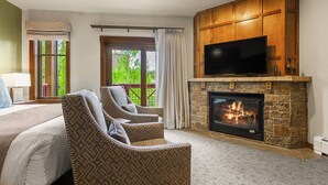 A bedroom with a large bed, two patterned armchairs, a fireplace, a TV, and a view of greenery outside. - Hyatt Main St Studio | Steps to Peak 9 + Downtown (Breckenridge)