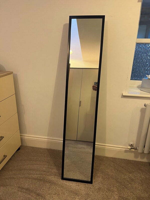 A tall, black-framed mirror in a room with beige walls, a white dresser, and a window with a view of a blue sky.