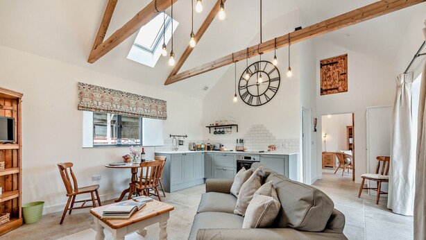 Ein geräumiges Wohnzimmer mit einer großen Couch, einem Holztisch und einem Essbereich mit Tisch und Stühlen. Im Hintergrund ist eine Küche mit einer großen Uhr und Pendelleuchten zu sehen.
