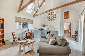 A spacious living room with a large sofa, a wooden coffee table, and a dining area with a table and chairs. A kitchen with a large clock and pendant lights is visible in the background. - Wonderful barn conversion Dog Friendly - Rabbit (Charlbury)