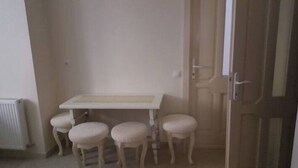 Una habitación pequeña y cerrada con una mesa blanca y cuatro sillas a juego. Se ve un radiador a la izquierda, y una puerta de madera a la derecha.