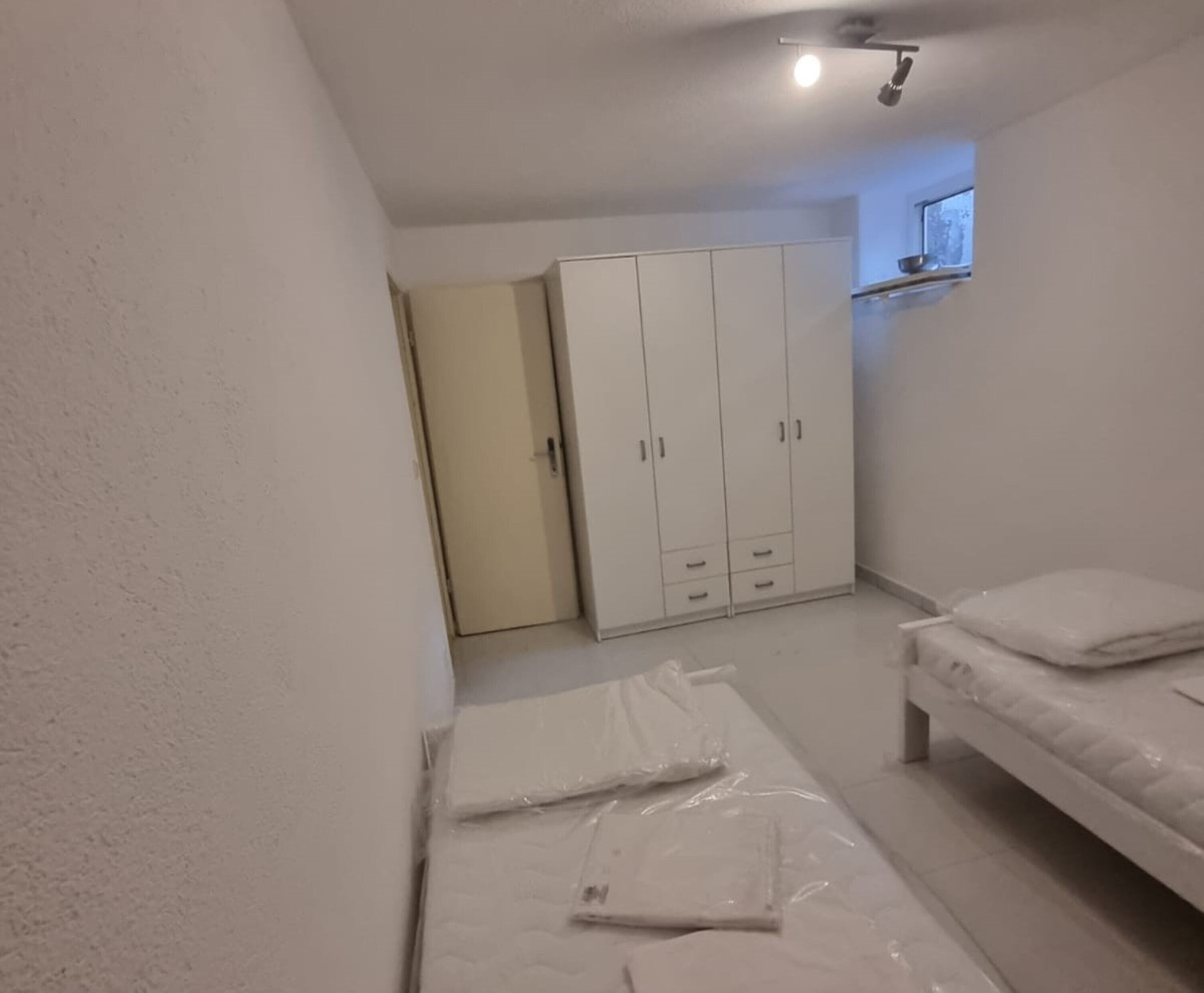A small, sparsely furnished room with a bed, a wardrobe, and a door leading to another room.