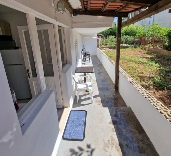 A covered outdoor area with a table and chairs, a door leading to a kitchen, and a small garden with a fence.