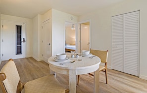 A dining area with a round table, two chairs, a bowl, a glass, and a spoon. There is a door with a window and a closet with louvered doors. - Stylish 1BR Condo · Walk to Kahana Beach (Lahaina)