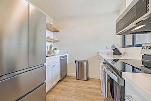 A modern kitchen with stainless steel appliances, wooden shelves, and a trash bin. - Stylish 1BR Condo · Walk to Kahana Beach (Lahaina)
