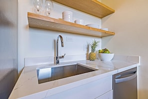 A kitchen with a stainless steel sink, wooden shelves above, and a bowl of fruit on the counter. - Stylish 1BR Condo · Walk to Kahana Beach (Lahaina)