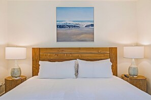 A bed with a wooden headboard, two bedside lamps, and a framed picture of a beach scene above it. - Stylish 1BR Condo · Walk to Kahana Beach (Lahaina)