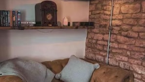 A cozy room with a brown leather sofa, two pillows, a bookshelf with books, a decorative clock, and a stone wall. - Charming and Modern 3 bed Cottage in Wansford (Peterborough)