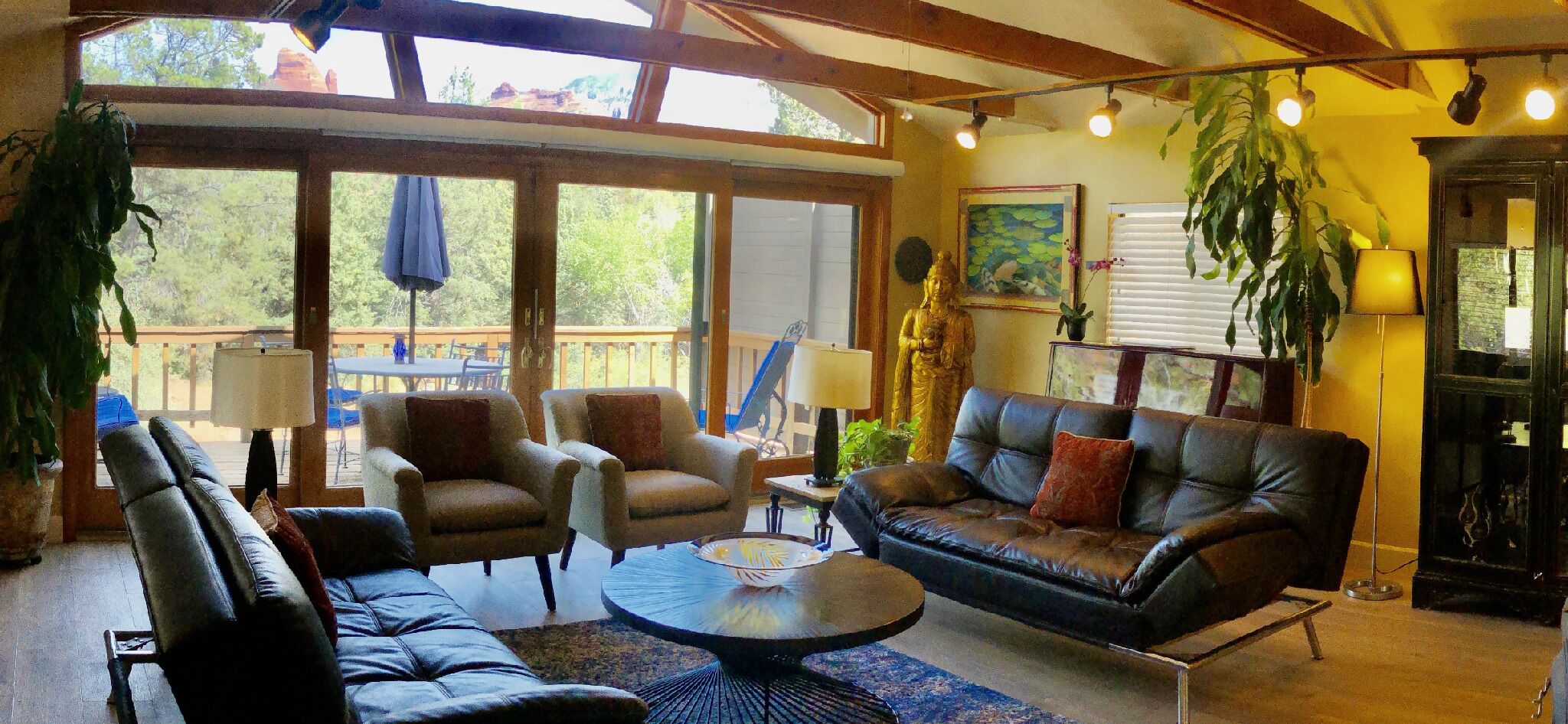 A cozy living room with a leather sofa, armchairs, and a coffee table. Large windows offer a view of the outdoors.