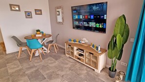 Ein Wohnzimmer mit einem an der Wand befestigten Flachbildfernseher, der verschiedene Streaming-Anwendungen anzeigt. Ein Holz-Unterhaltungsmöbel mit Stauraumregalen und einem Blumentopf sind vorhanden. Im Hintergrund ist ein Essbereich mit Tisch und Stühlen zu sehen.