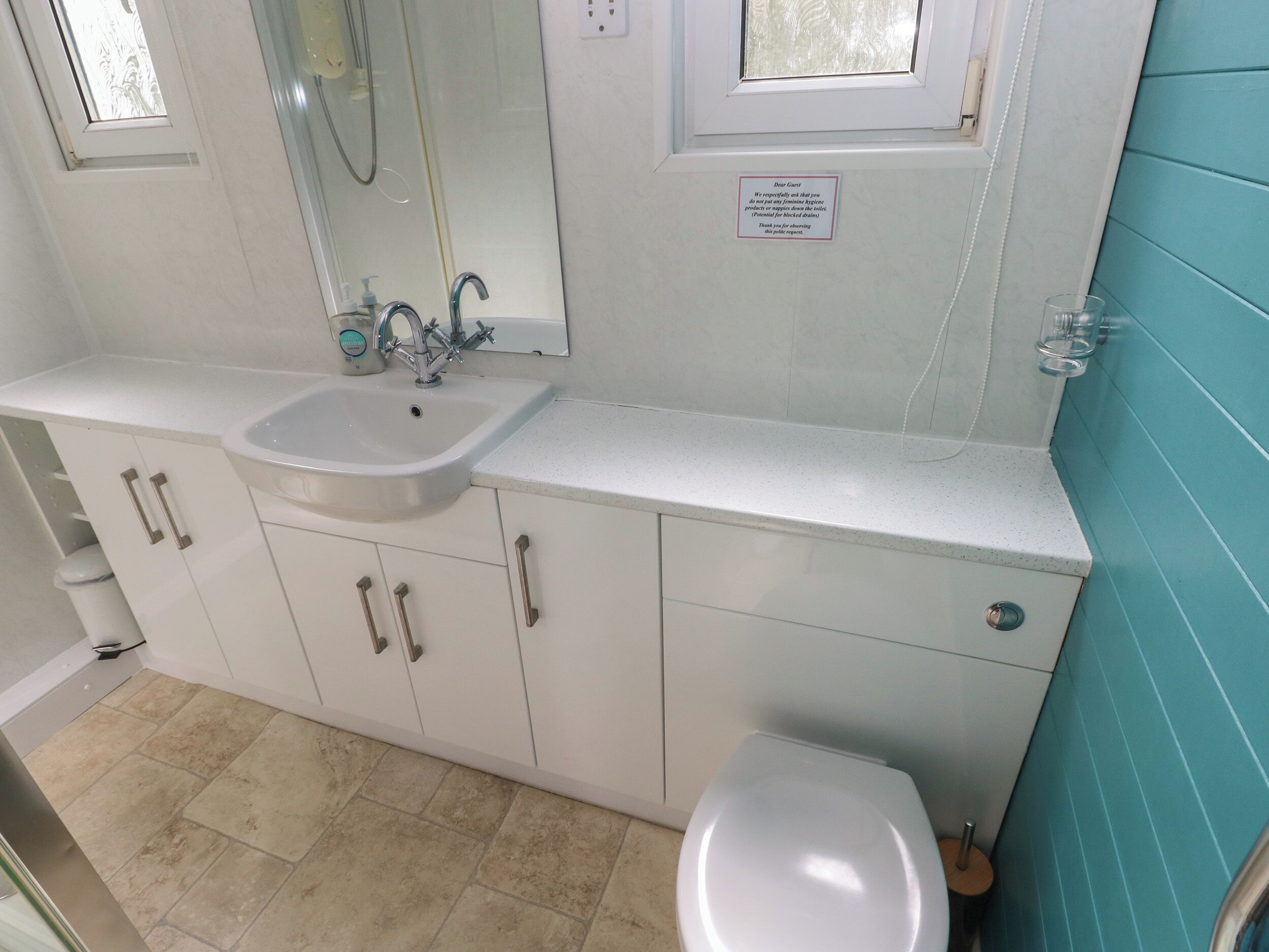 A bathroom with a white sink, mirror, and toilet. There is a small window above the sink and a sign on the wall.