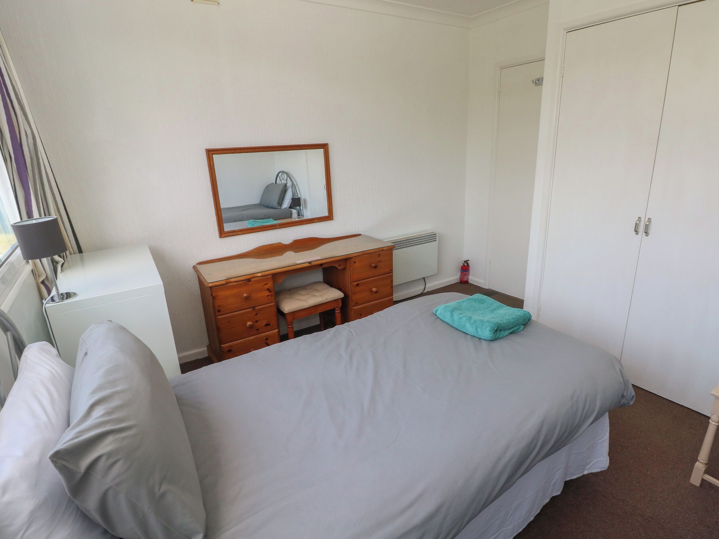 A bedroom with a bed, a wooden dresser, a desk, a chair, a mirror, a radiator, and a wardrobe.