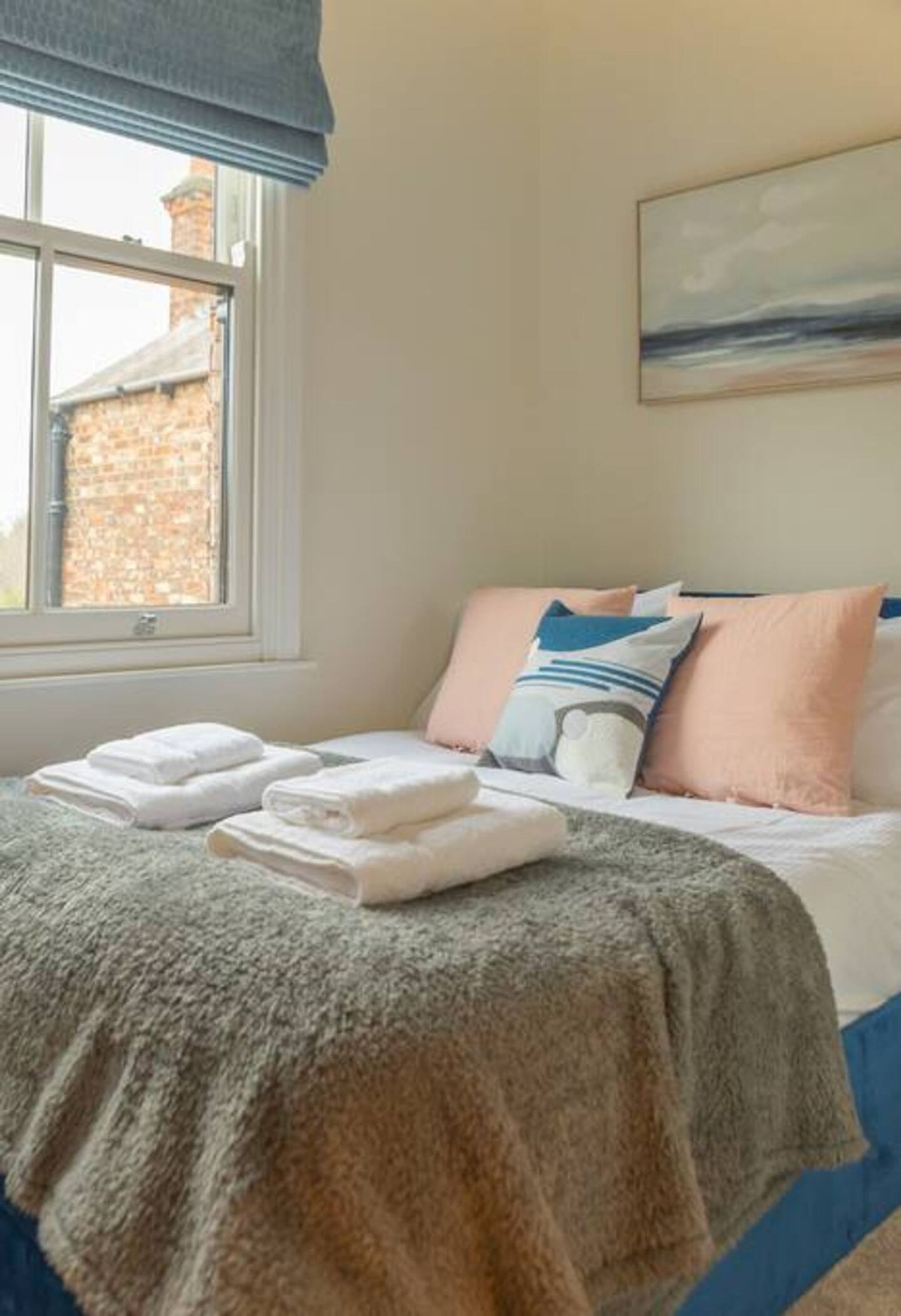 Una cama bien hecha con almohadas y una manta, una ventana con vista a un edificio de ladrillo, y un cuadro en la pared.