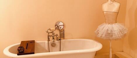 A bathroom with a clawfoot bathtub, a white vanity, and a mannequin dressed in a tutu.