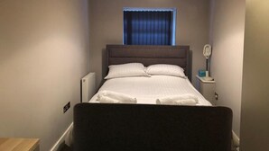 Un lit bien fait dans une petite pièce avec une fenêtre, une table de chevet avec une lampe et un radiateur.