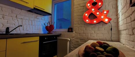A kitchen with yellow cabinets, a wooden countertop, a bowl of fruit, and a neon sign.