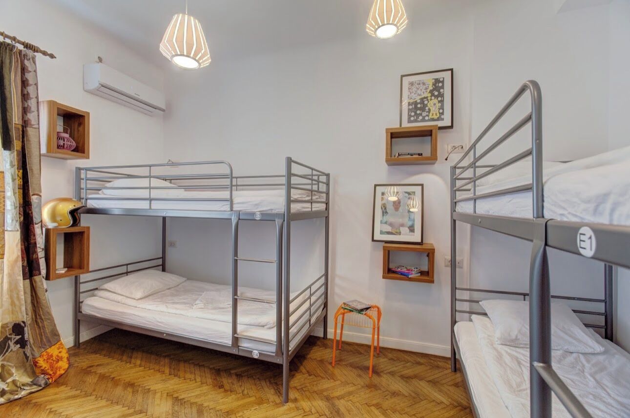 Habitación con literas, suelo de madera, un taburete pequeño y una estantería con libros.