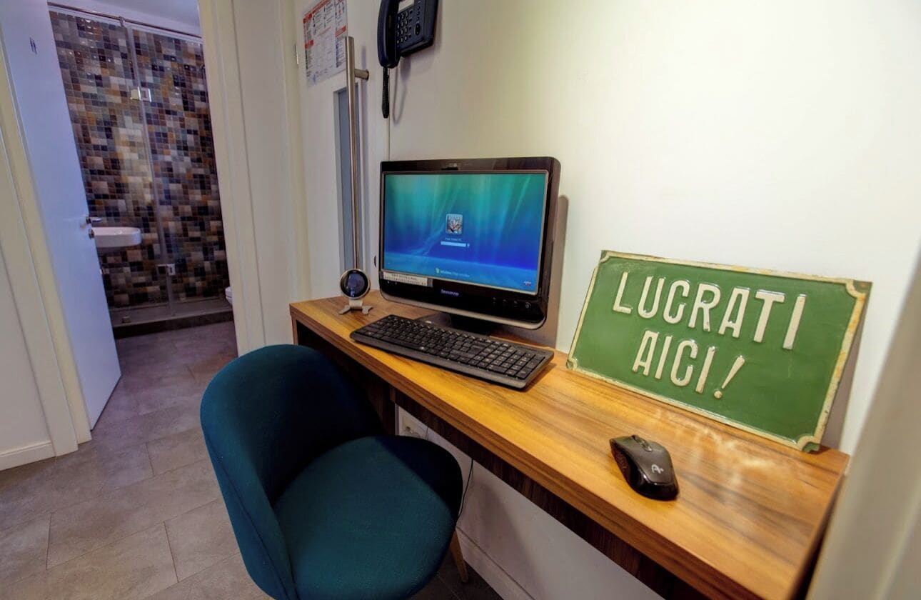Una oficina pequeña con una computadora de escritorio, un teclado, un ratón y un cartel que dice "LUCRATI AICI!".