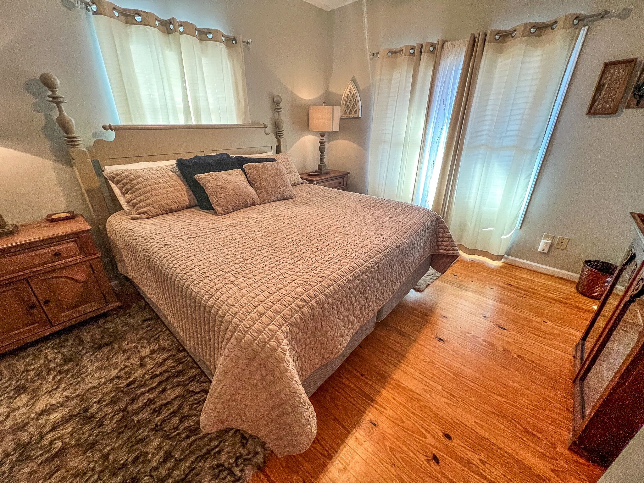 Un dormitorio con una cama grande, cabecera de madera, una mesita de noche, una lámpara y una ventana con cortinas.
