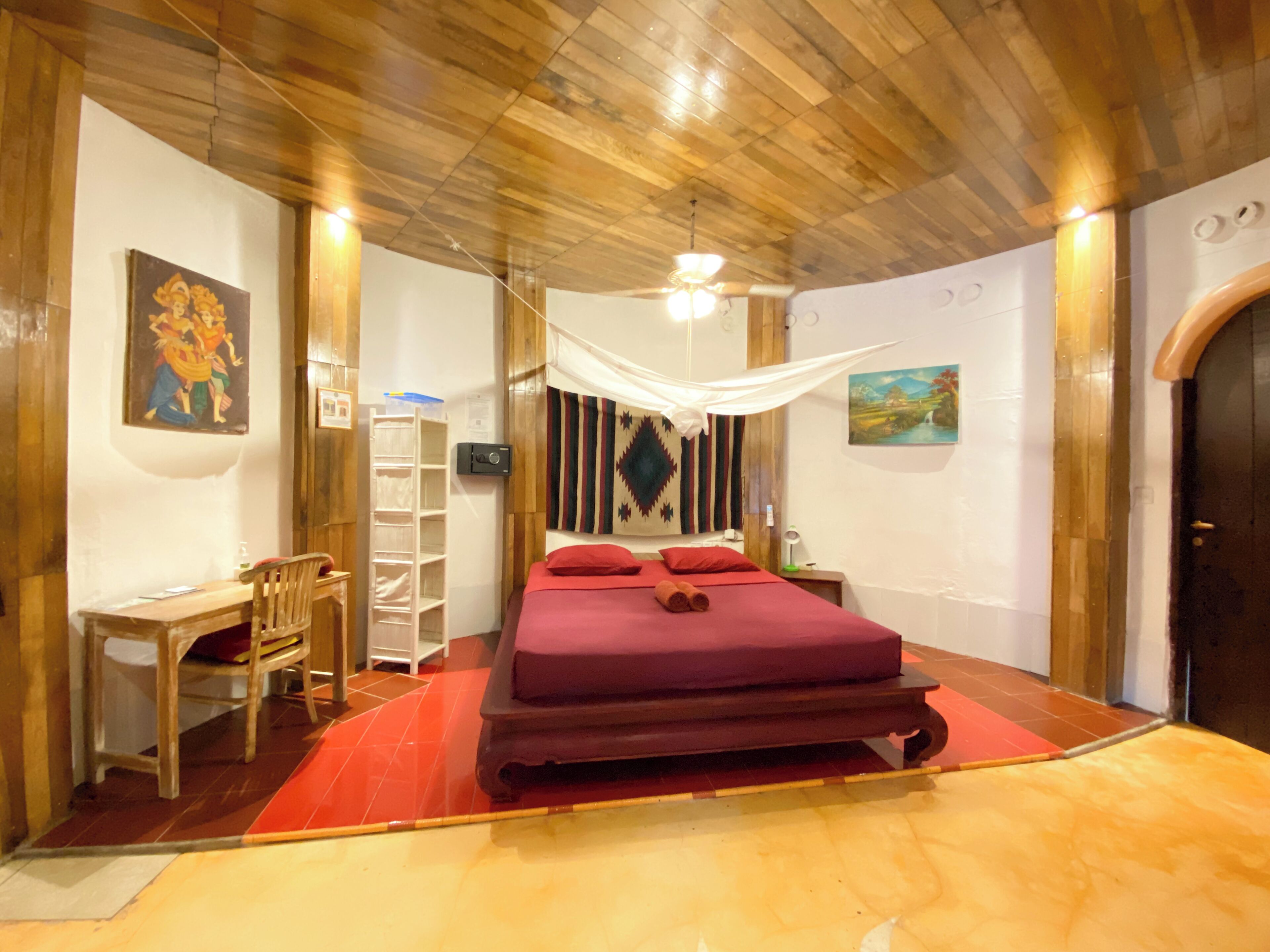 Una habitación con techo de madera, una cama con cubrecama rojo, un escritorio con silla y una estantería.