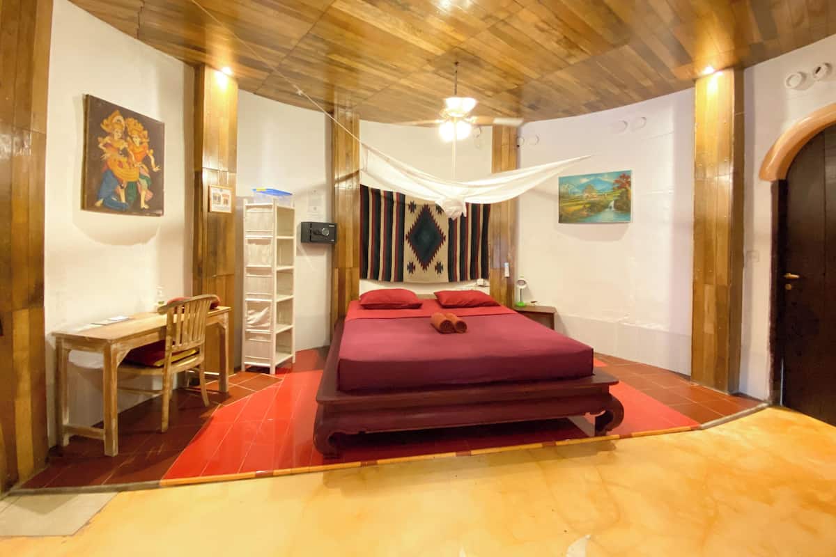 A room with a wooden ceiling, a bed with a red bedspread, a desk with a chair, and a shelf unit.