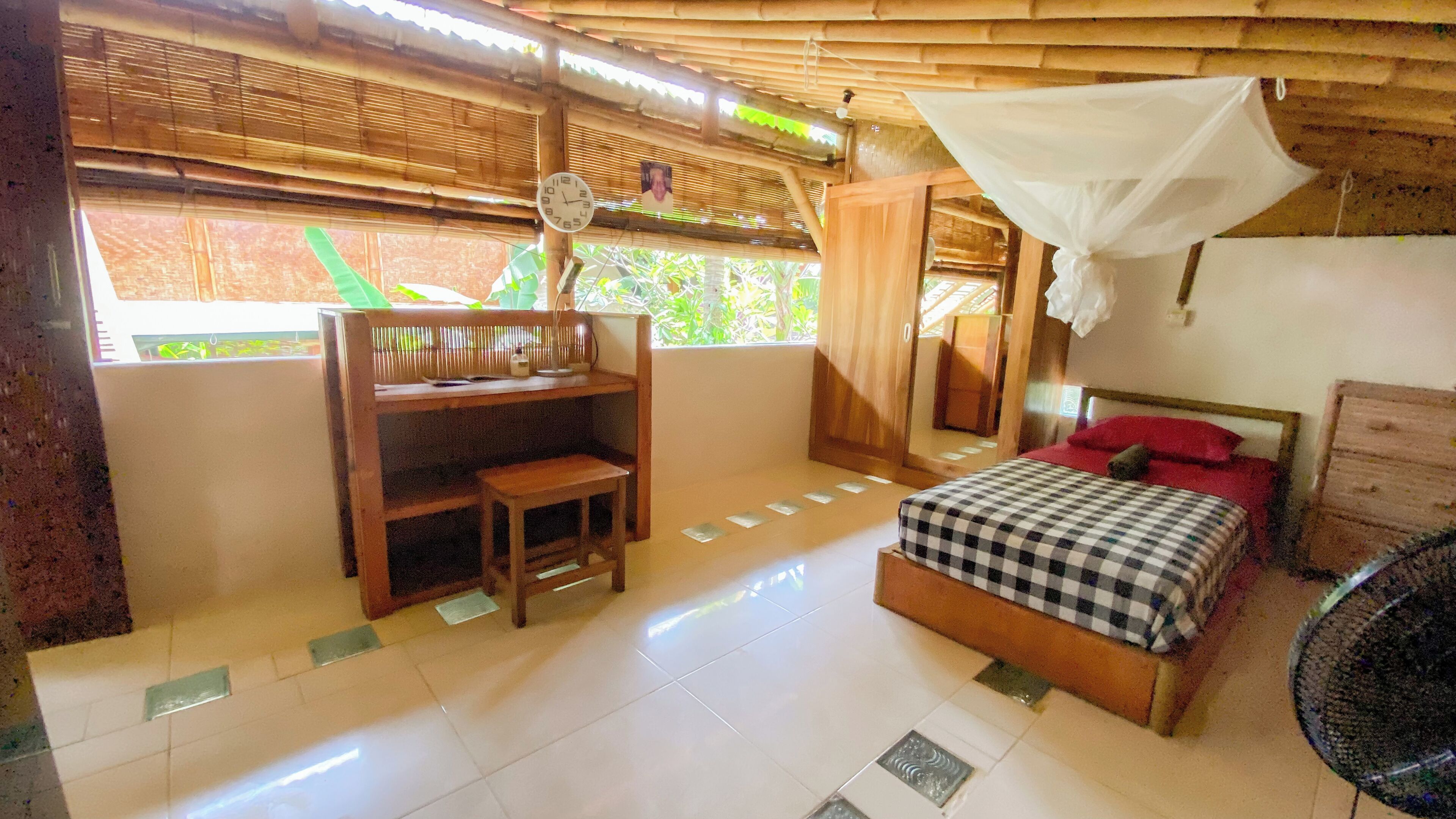 Una habitación con cama, escritorio, silla, mesita de noche, ventilador y ventana con vistas a zonas verdes.